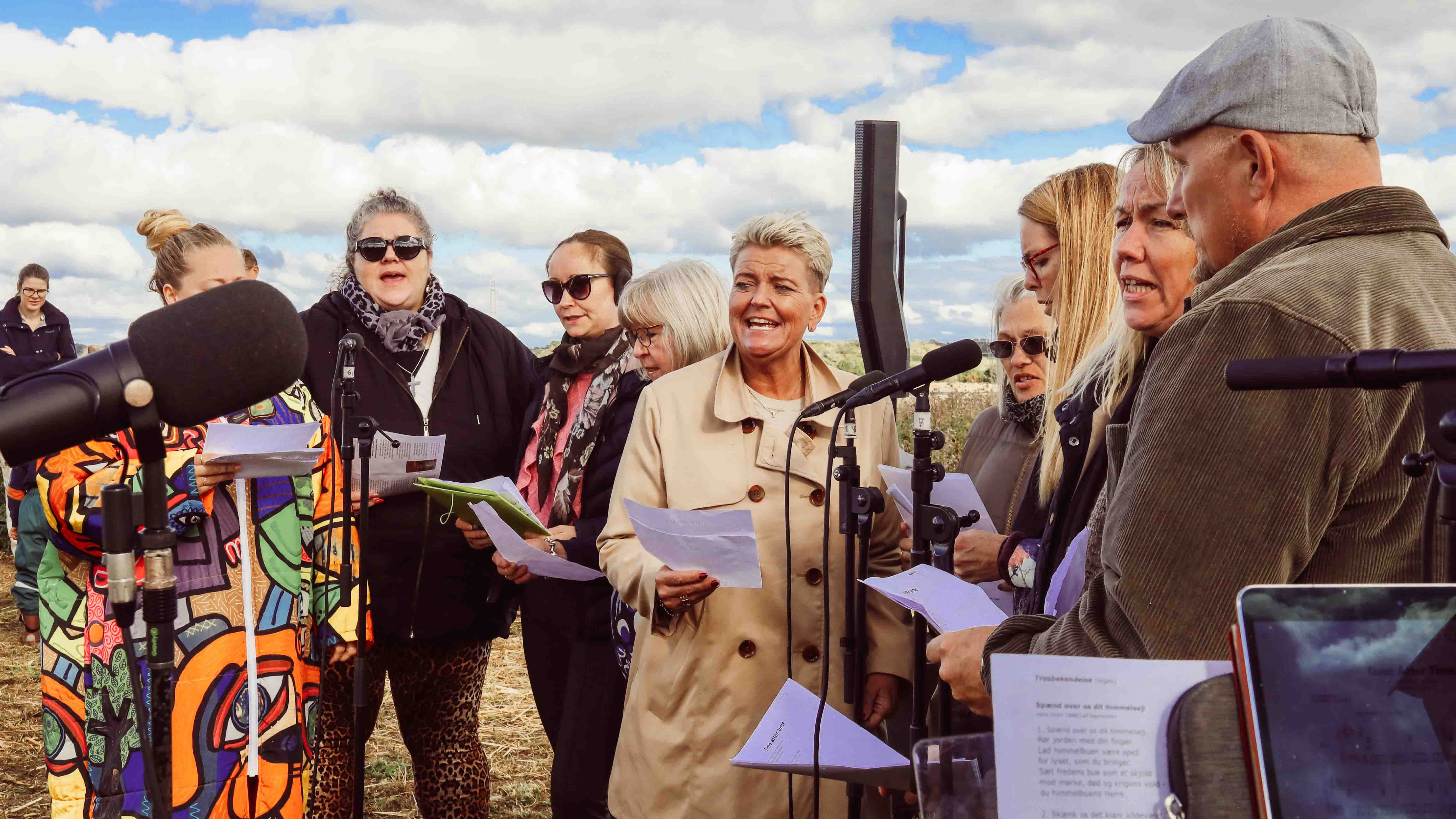 Koret PopShop i Ølstykke Sogn synger til udendørs høstgudstjeneste på Herrens Mark september 2025. Korsangere med noder og mikrofoner synger.